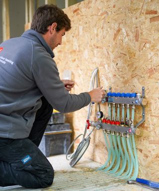 Man installing a JK Floorheating's underfloor heating manifold against a chipboard stud wall in a newly renovated house