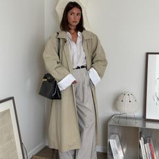 A woman wears a trench coat, button down shirt, trousers, and black leather hermes bag.