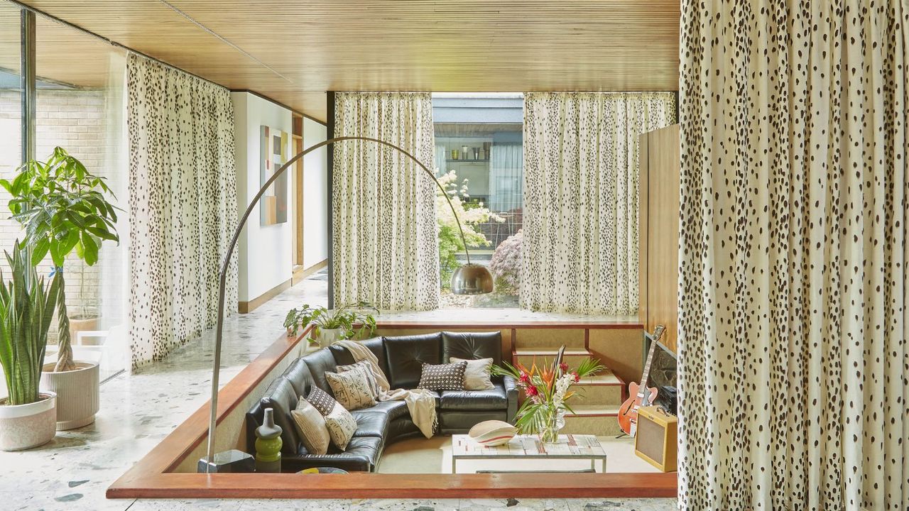 A living room with windows with white and brown polka dot curtains around them, white marble tiling, and a lowered seating area with a gray leather couch, a coffee table, and a guitar in it.