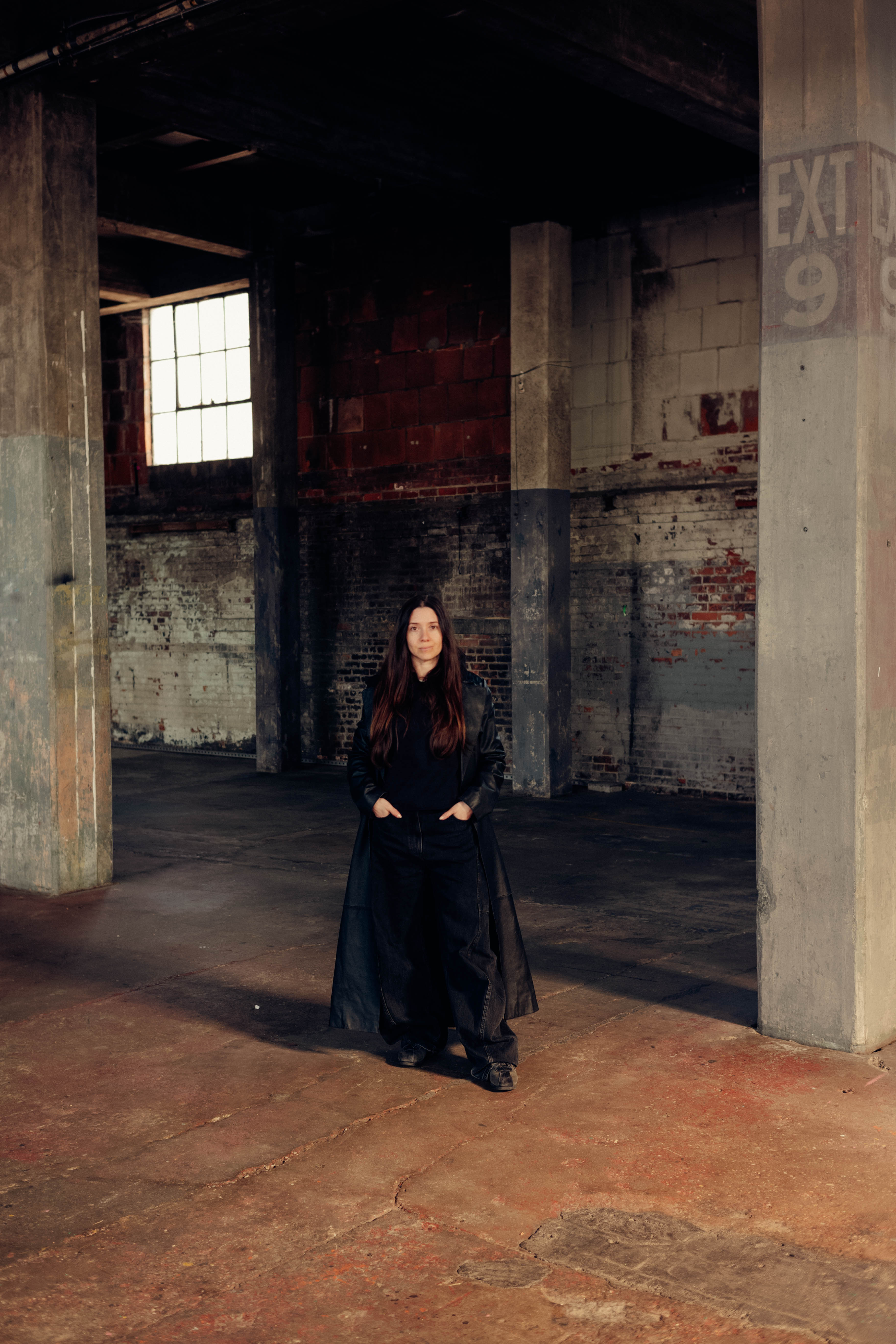 A woman dressed in a full black outfit, including a leather coat, black trousers, and top, stands with her hands in her pockets in a bare industrial site.