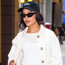 Tessa thomspon wears a black baseball cap, sunglasses, white tee, and a white barn jacket.