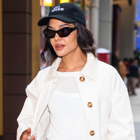 Tessa thomspon wears a black baseball cap, sunglasses, white tee, and a white barn jacket.