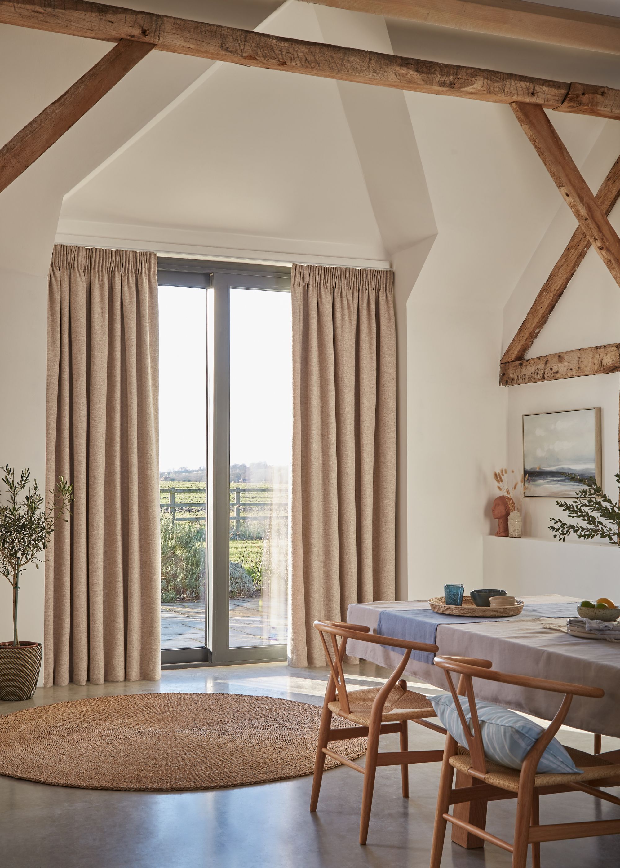 A modern rustic dining space with warm white walls and wood ceiling beams, sliding glass doors with beige curtains.