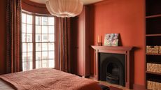A bedroom painted a terracotta red with a red and white striped headboard, white and brick red bedding, a nightstand with a wavy lampshade. terracotta bedroom