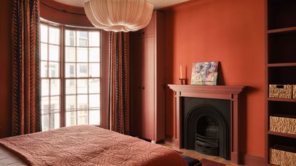 A bedroom painted a terracotta red with a red and white striped headboard, white and brick red bedding, a nightstand with a wavy lampshade. terracotta bedroom