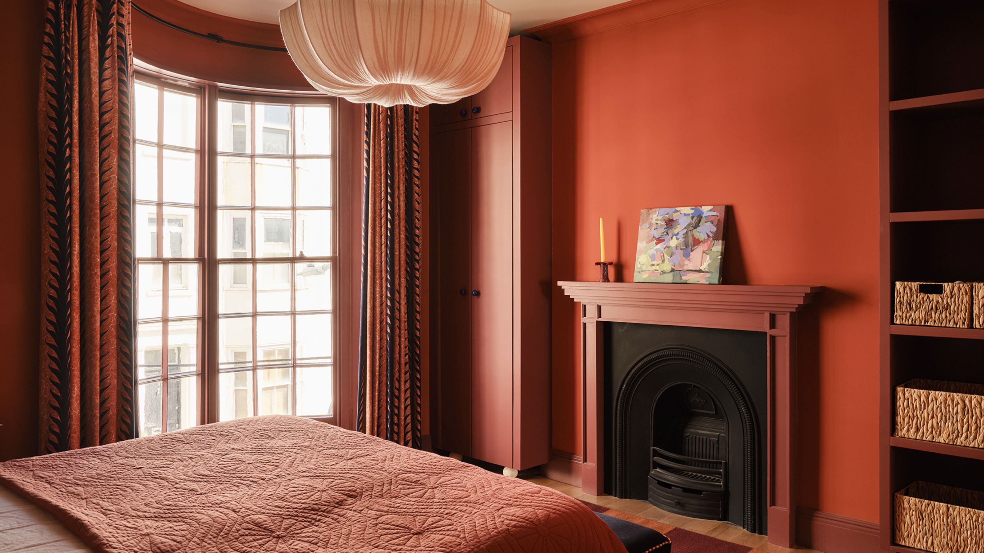 A bedroom painted a terracotta red with a red and white striped headboard, white and brick red bedding, a nightstand with a wavy lampshade. terracotta bedroom
