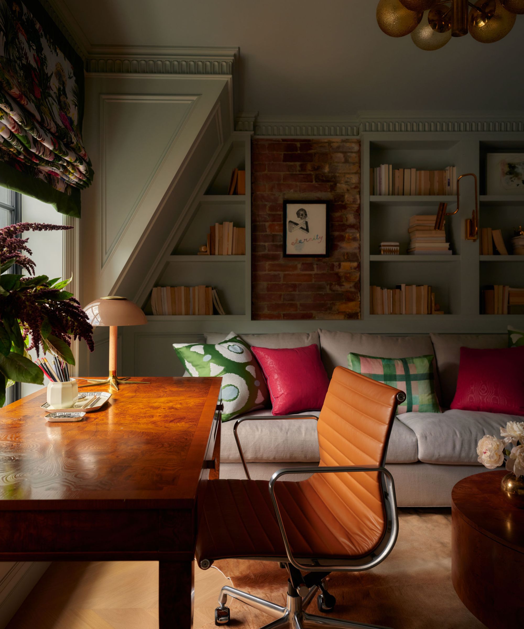 a home office with exposed brick and slanted ceilings, painted bookcases, a large couch and an antique wooden desk with tan leather chair
