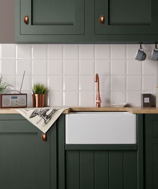 A kitchen with dark green cabinets with copper handles, a white tiled splashback, a copper faucet and a white ceramic basin