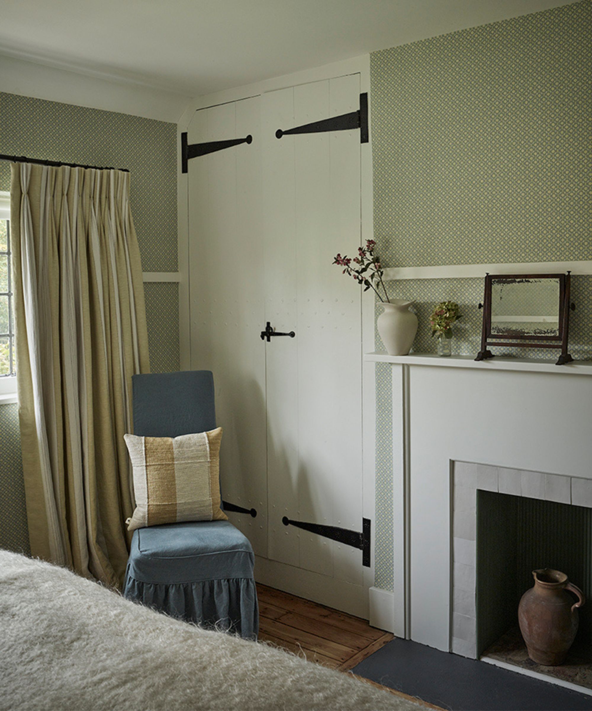 a corner of a cottage bedroom with green wallpaper, a restored fireplace and cupboard with a blue slipper chair styled with a yellow gingham pillow