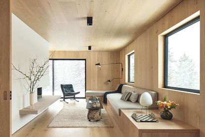 A view of a modern cabin style living space with sleek light wood floors, walls and ceiling, with large black framed windows, and a bench style built in sofa in a grey/beige fabric. 