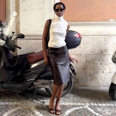 Paris-based fashion influencer Sylvie Mus poses on a street in Roma, Italy in front of a peach-colored wall and a motorcycle wearing black cat-eye sunglasses, a white sleeveless turtleneck top, shoulder bag, leather skirt, and strappy mule heeled slide sandals