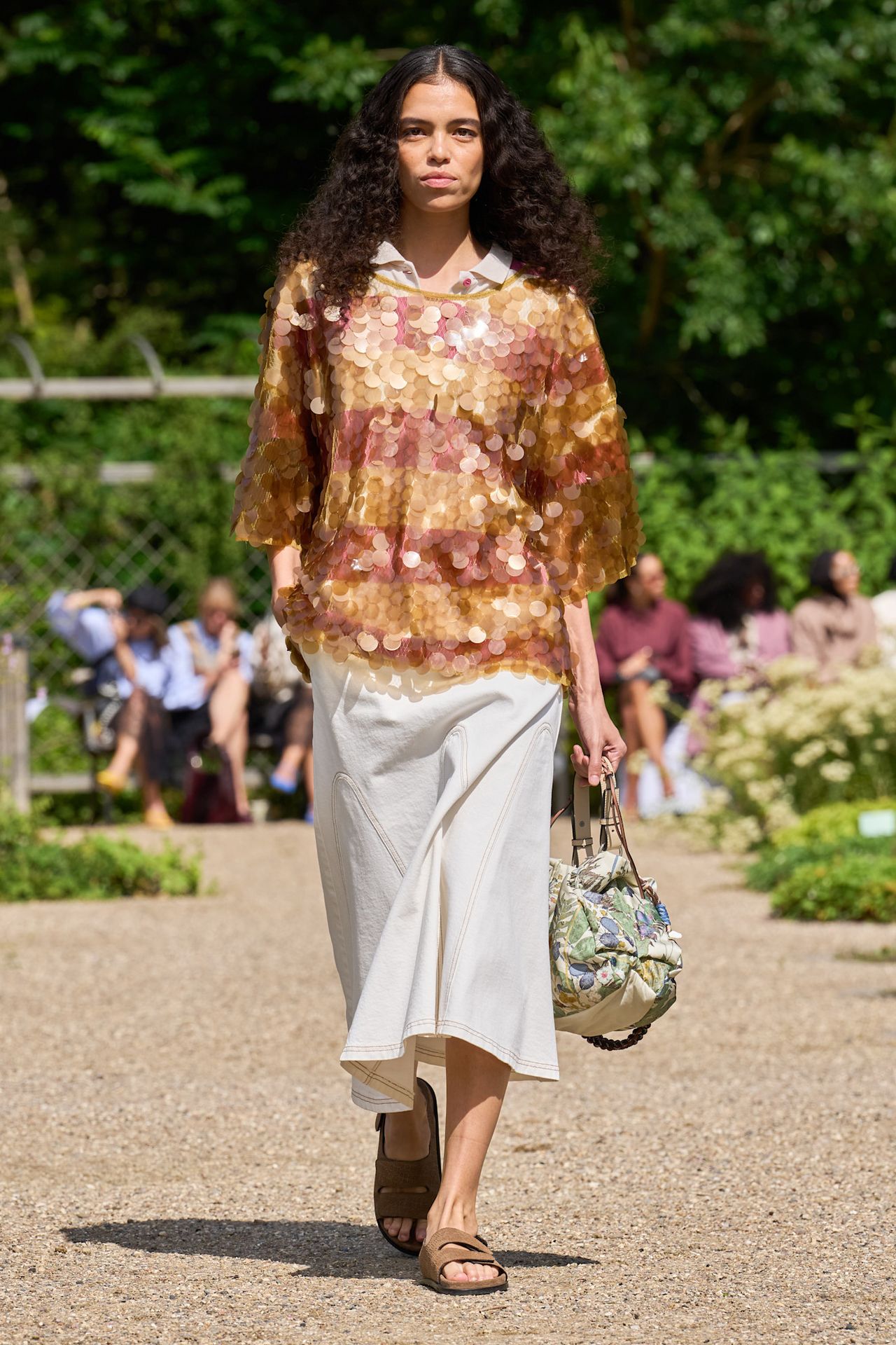 Layering staples for spring 2026 are shown in a photo from Munthe's runway show, with a model walking while wearing a tan polo shirt underneath a striped yellow and red sequin top styled with a white skirt, tan suede sandals, and a bucket bag