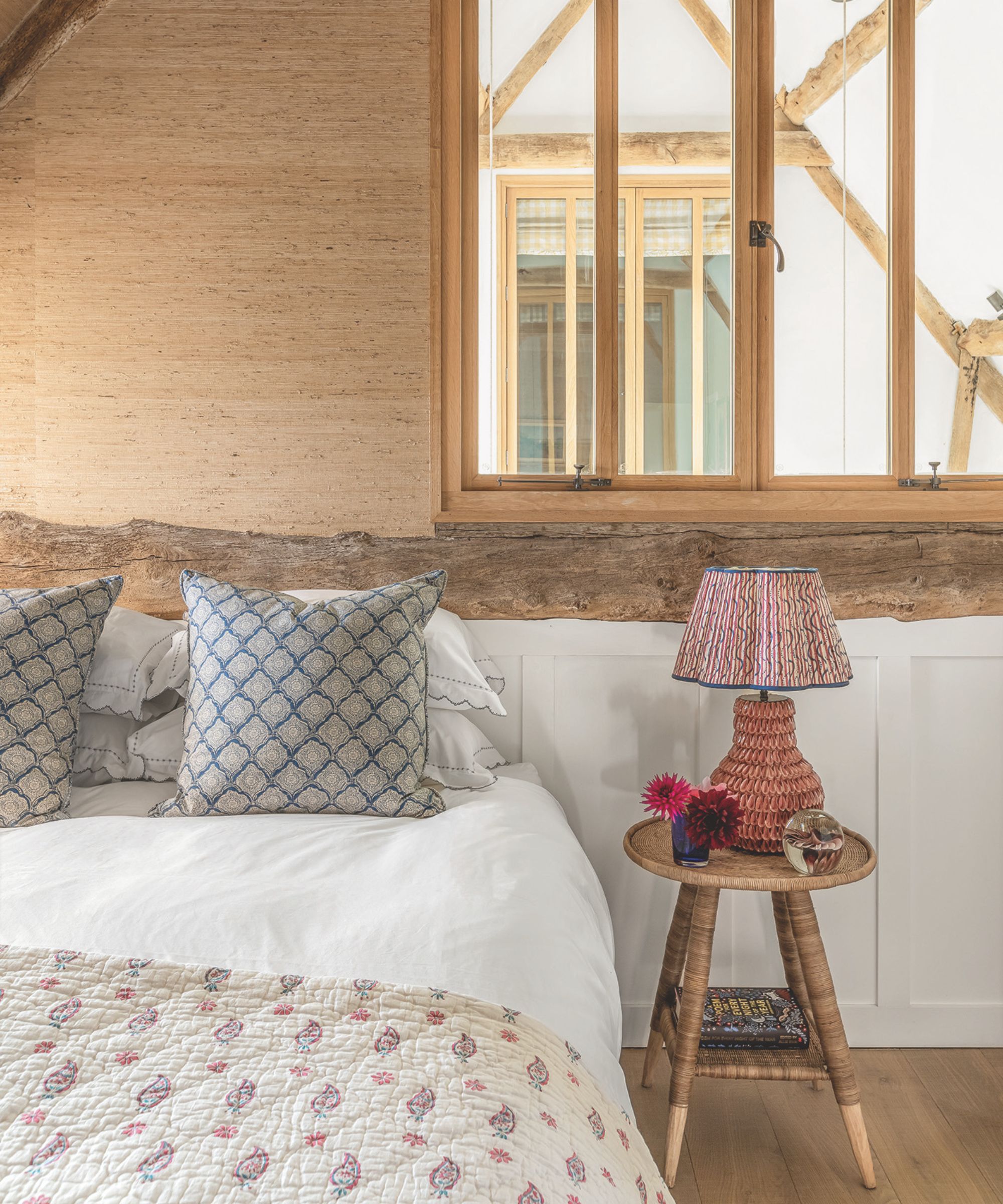 A wooden panelled attic bedroom space, with a double bed to the left with white bedding, patterned cushions, and a cream patterned quilt, and a circular bedside table to the right with a pink, patterned lamp, flowers, and a book. Above the bed, to the right, are large windows, and the wall above is wood.