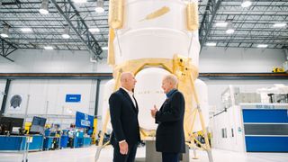two men in suits stand in a brightly-lit hangar in front of a capsule-shaped moon lander