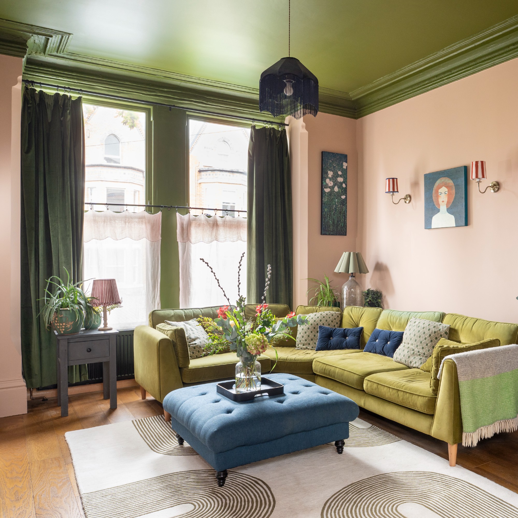 A pink living room with a contrasting green ceiling and a tonal green corner sofa with a blue ottoman used as a coffee table