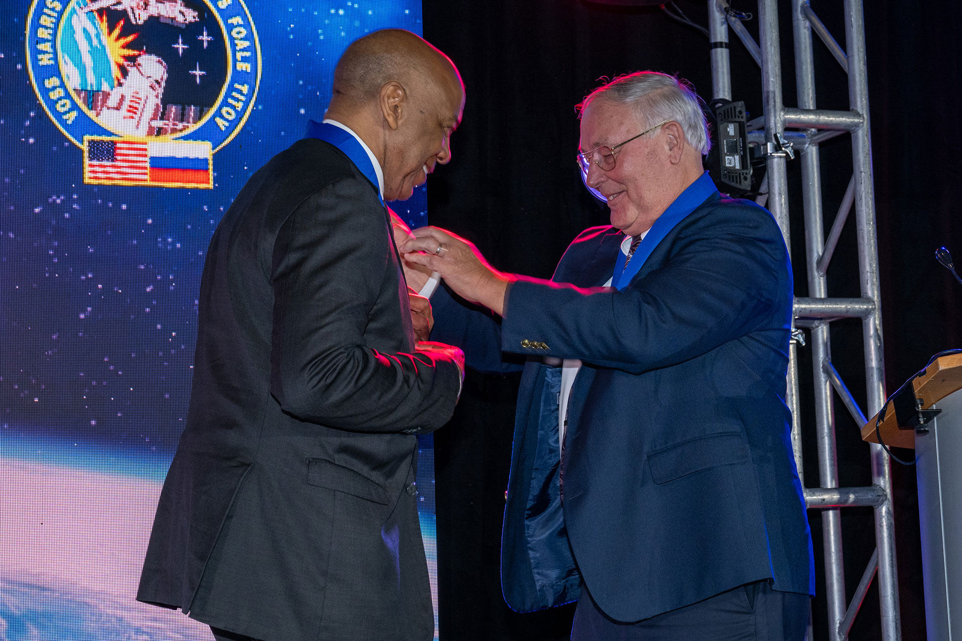 two men in dark color suits meet on stage, where one presents another a medal