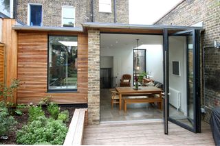a glass and wood extension attached to a home by IQ glass, with a wooden ding table and a tiled floor, leading out onto wood decking
