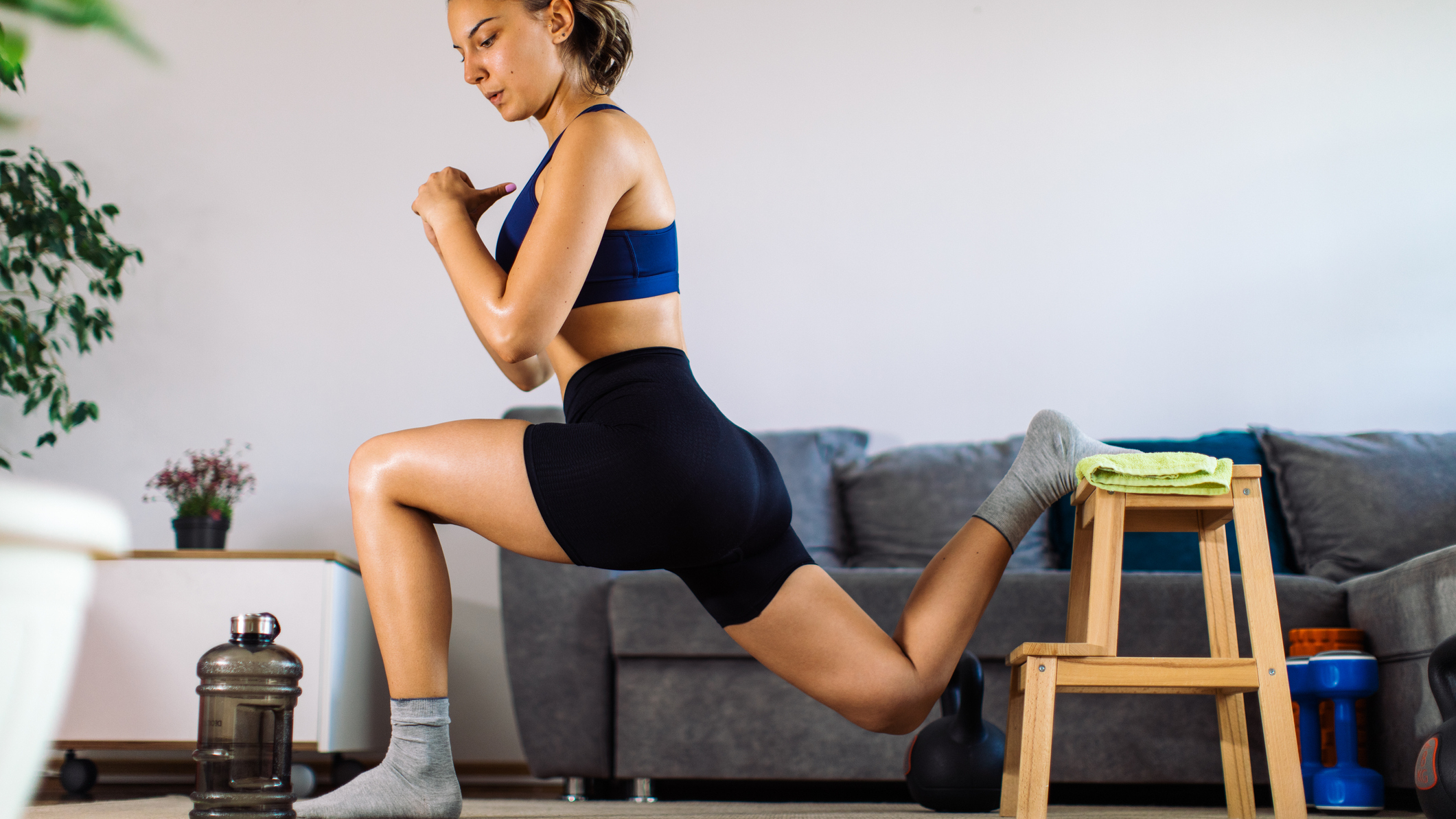 Woman performs Bulgarian split squat at home with her rear foot elevated on a foot stool