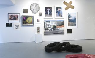Art gallery featuring grey floors and white walls with framed art displays in different sizes. 3 black tyre -like objects placed on each other in the middle of the room.