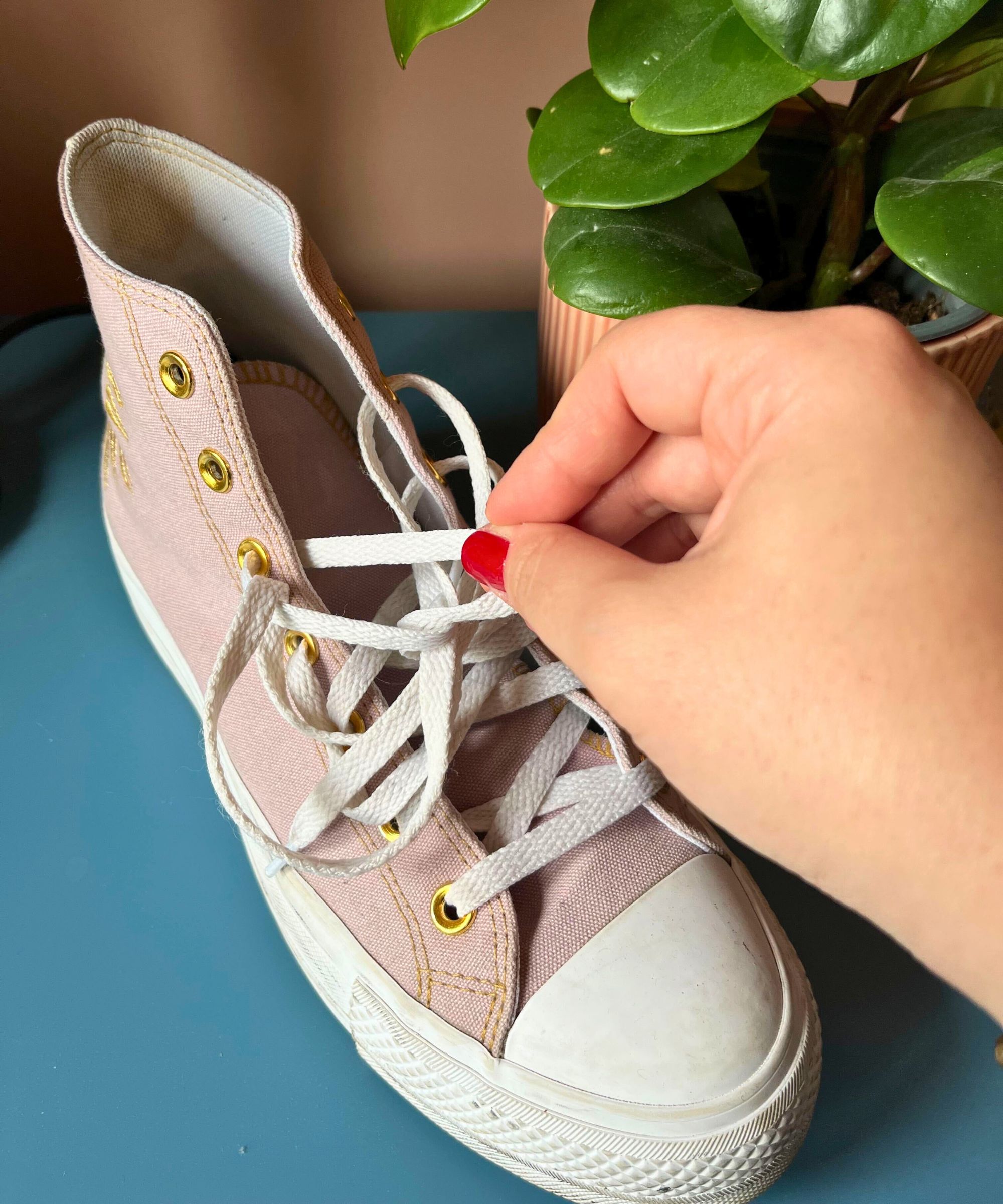 A lilac pair of high-top Converse shoes on a blue counter, with a plant visible in the background. There is a hand in shot, pulling out the white laces