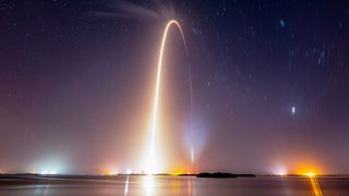 a time lapse view of a night time launch showing the arc of the rocket reflecting in body of water in the foreground