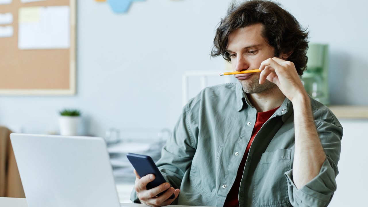 A young man plays with a pencil while looking at his phone and laptop.