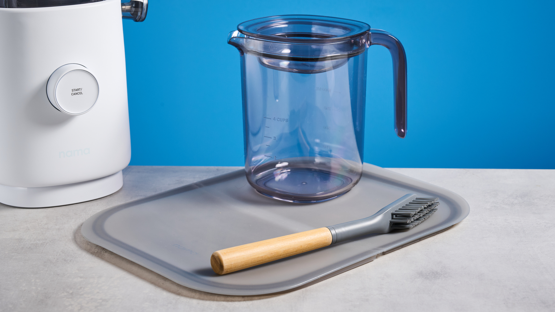 the nama m1 plant-based milk maker in white with a pitcher, blade frame, and strainer, and a pulp mat accessory with a brush