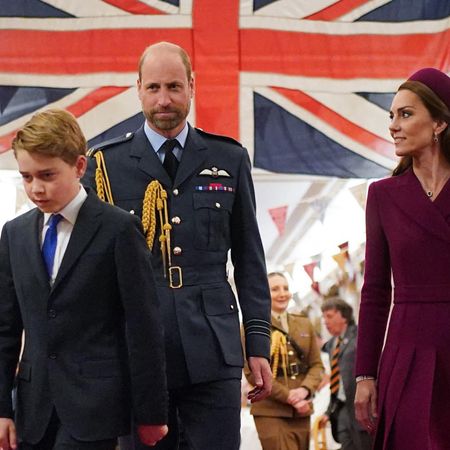 Prince George wears a navy suit and walks in front of a large Union Jack flag with his parents, Prince William in a navy military uniform and Princess Kate in a dark red coat dress