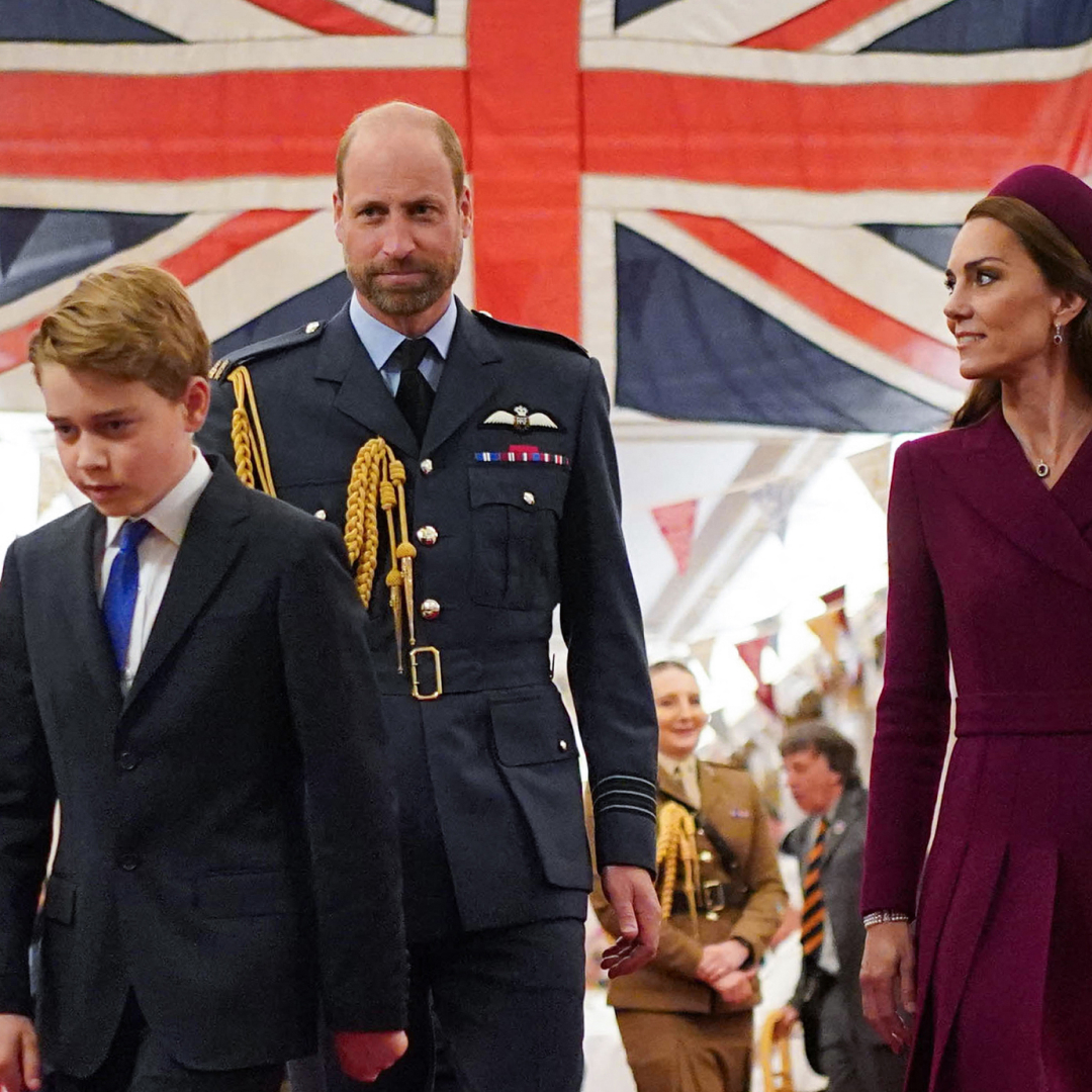 Prince George wears a navy suit and walks in front of a large Union Jack flag with his parents, Prince William in a navy military uniform and Princess Kate in a dark red coat dress