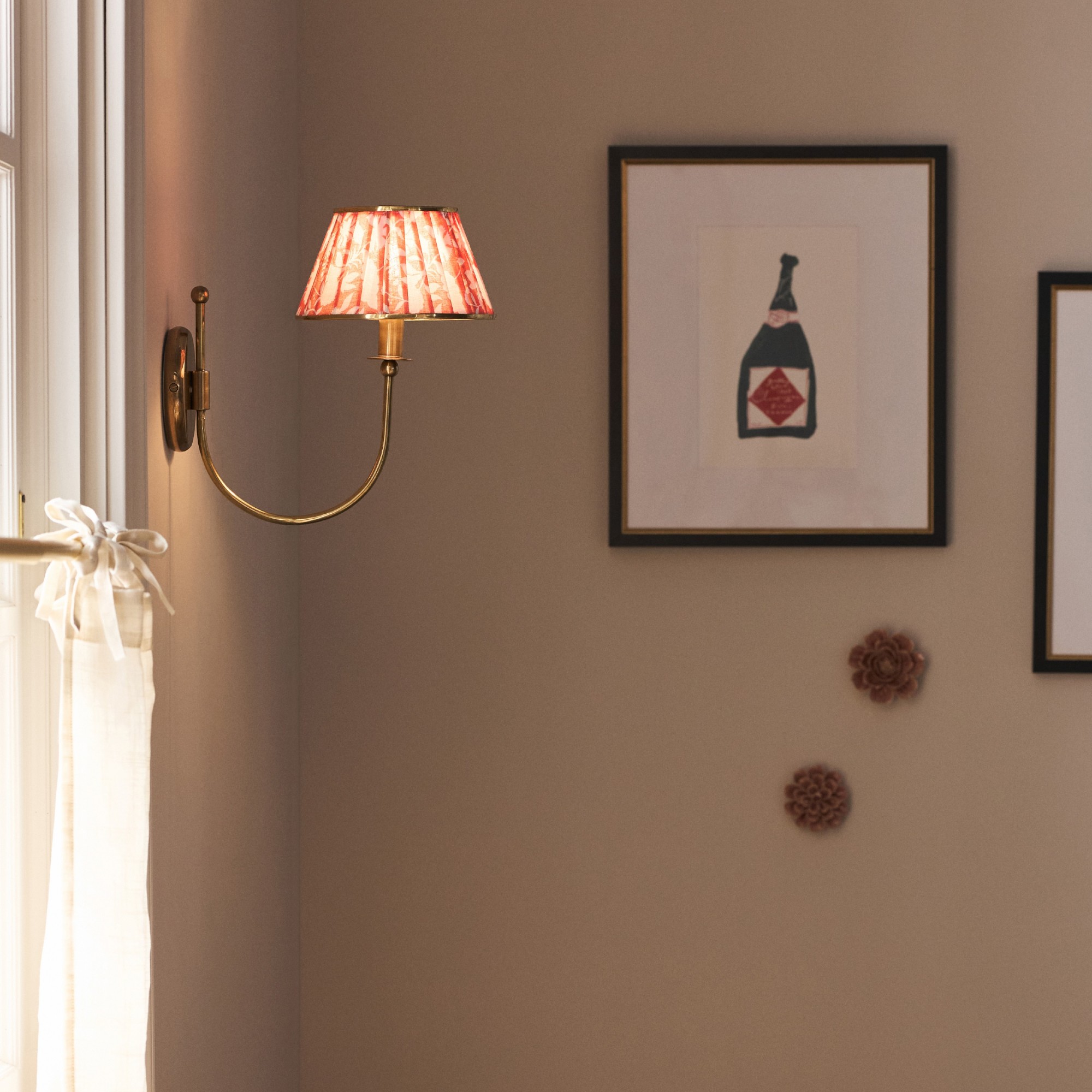 A switched-on wall light from Anthropologie next to a window with a cafe curtain and framed art print in the background