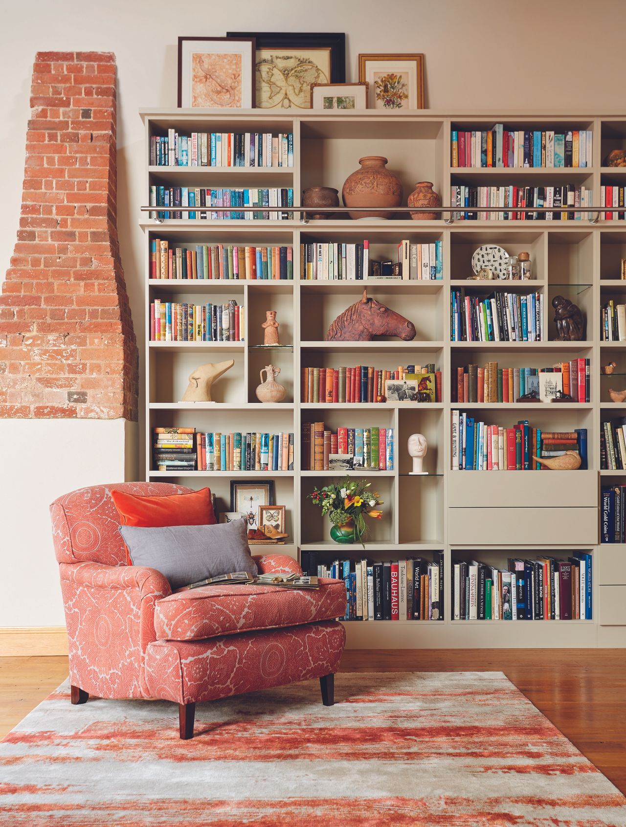 a luxurious spare room with a red armchair and rug, and a huge sage green built in bookcase unit with lots of books in it, and a brick covered-up fireplace next to it