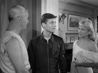 a still from a black and white twilight zone sketch of a man looking confused in a couple's living room