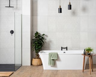 Freestanding white bath with white tiled wall, shower unit and indoor houseplant on wooden flooring
