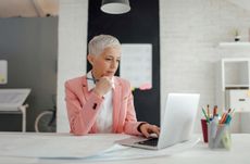 A woman with silver hair in a pink suit jacket looks at a laptop on a work desk with a cup of coffee and writing utensils in a sleek office.