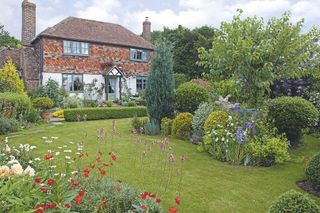 17th century cottage garden with a neat manicured lawn and flowering sections of gardens with trees, and a traditional Tudor house beyond