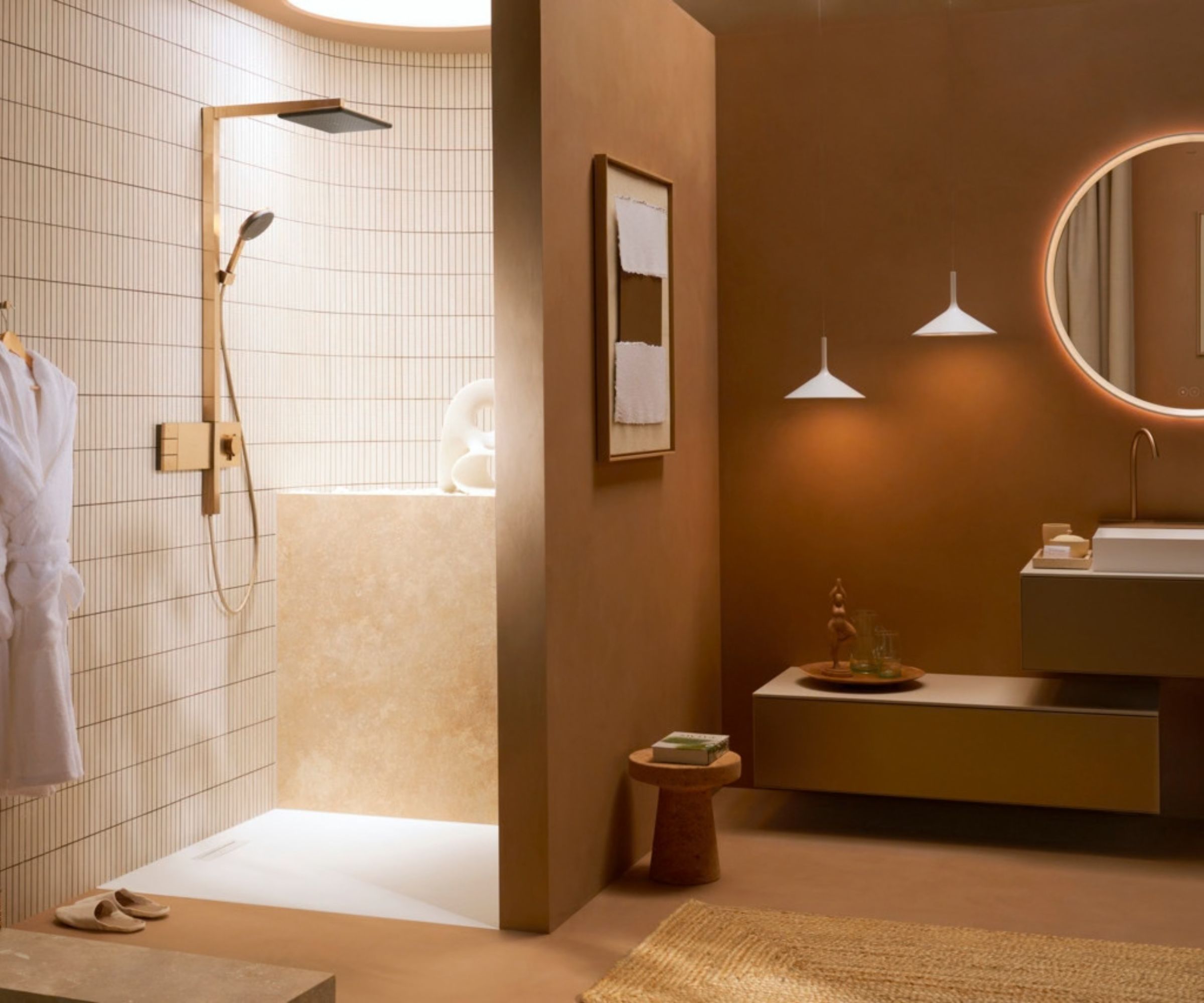A warm, terracotta-toned bathroom with a walk-in shower featuring vertical white tiles and brass fittings. The vanity area has deep brown walls, a floating unit with a white rectangular basin, a backlit round mirror and two white cone pendant lights.