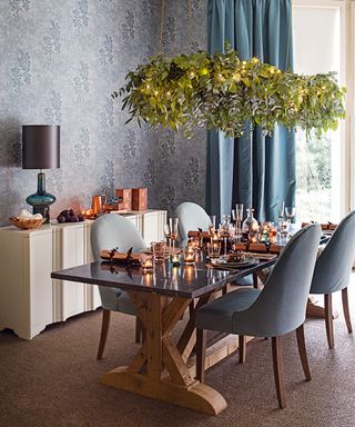 Blue dining room with wooden table and blue fabric covered chairs. Foliage pendant light. Blue patterned wall paper. Blue curtains.