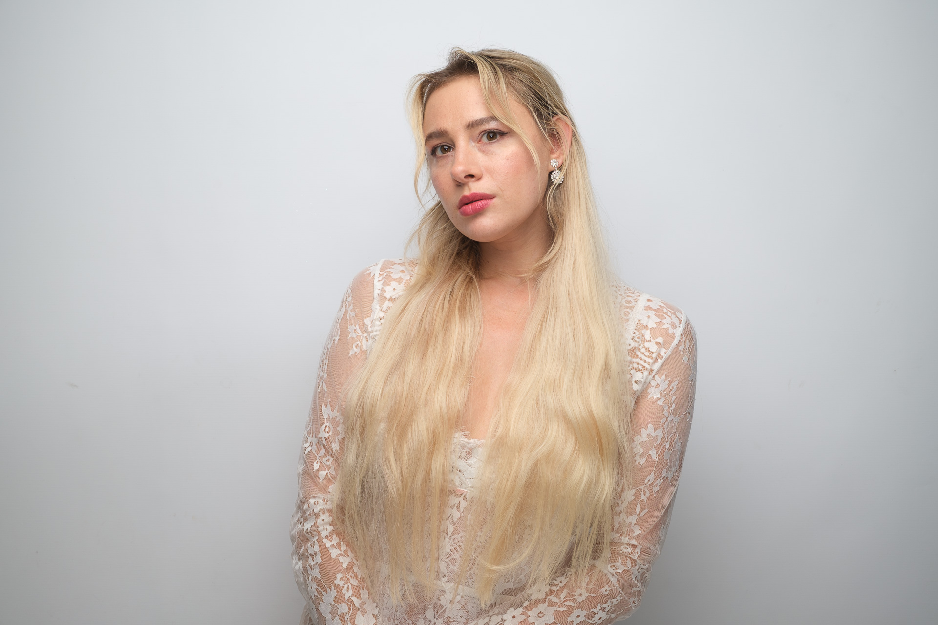 Portrait of a blonde woman wearing a white lace outfit and jeweled earrings against a plain light-gray background.