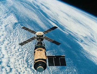 a large space station above earth with four large solar panels in a windmill type orientation and a long cylindrical body.