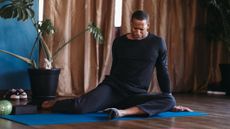 Man on yoga mat stretching out hips; behind him is a leafy plant and massage ball