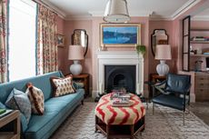 A small living room with pink walls, blue sofa and red and white patterned ottoman, drapes and cushions.
