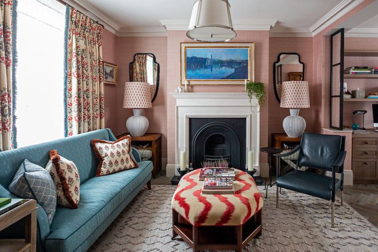 一个带粉红色墙壁，蓝色沙发和红色和白色图案无背长椅，窗帘和靠垫的小型客厅。