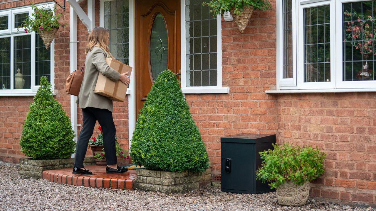 Yale Smart Delivery Box review a black Yale delivery box beside the front door of a house beside plant pots while a woman walks in the front door with a package