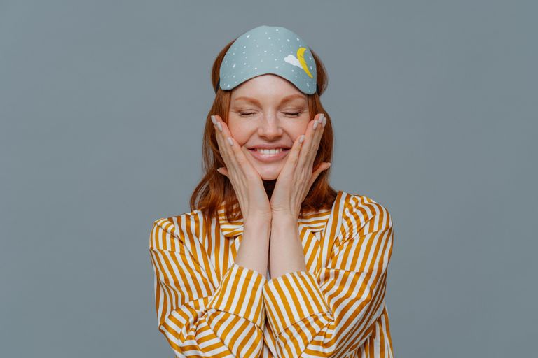 Smiling Woman Wearing Eye Mask With Eyes Closed Standing Against Gray Background - Best Sleep Aid