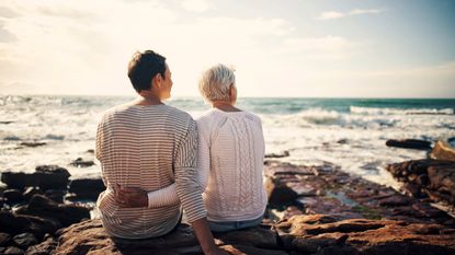 Rear view of senior woman and adult daughter sitting on rocks at the beach and looking out to sea