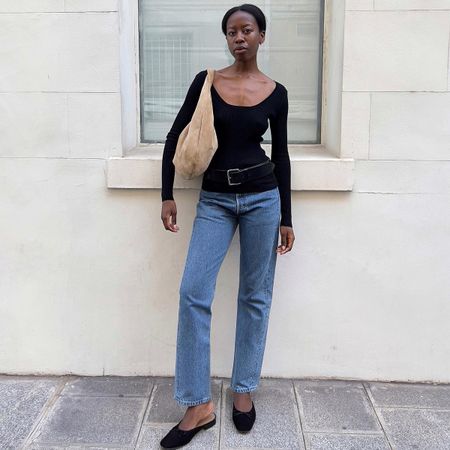 Sylvie Mus wears a black long-sleeve top, blue jeans, black belt, black mules, and a beige suede bag.