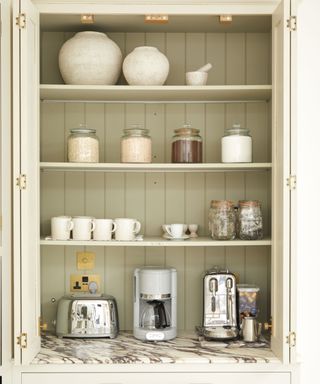 A compact pantry cupboard with sage green vertical panelled backing, featuring open shelving. The top shelf holds two textured ceramic vases and a mortar and pestle. Below are jars filled with dry goods, a row of mugs, a stainless steel toaster, a coffee machine, and an espresso maker on a marbled countertop.