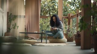 Former RA MA employee Nicole Norton, looking at her phone as she sits cross-legged on a floor pillow in a room with glass walls and trees outside, in 'Breath of Fire.'