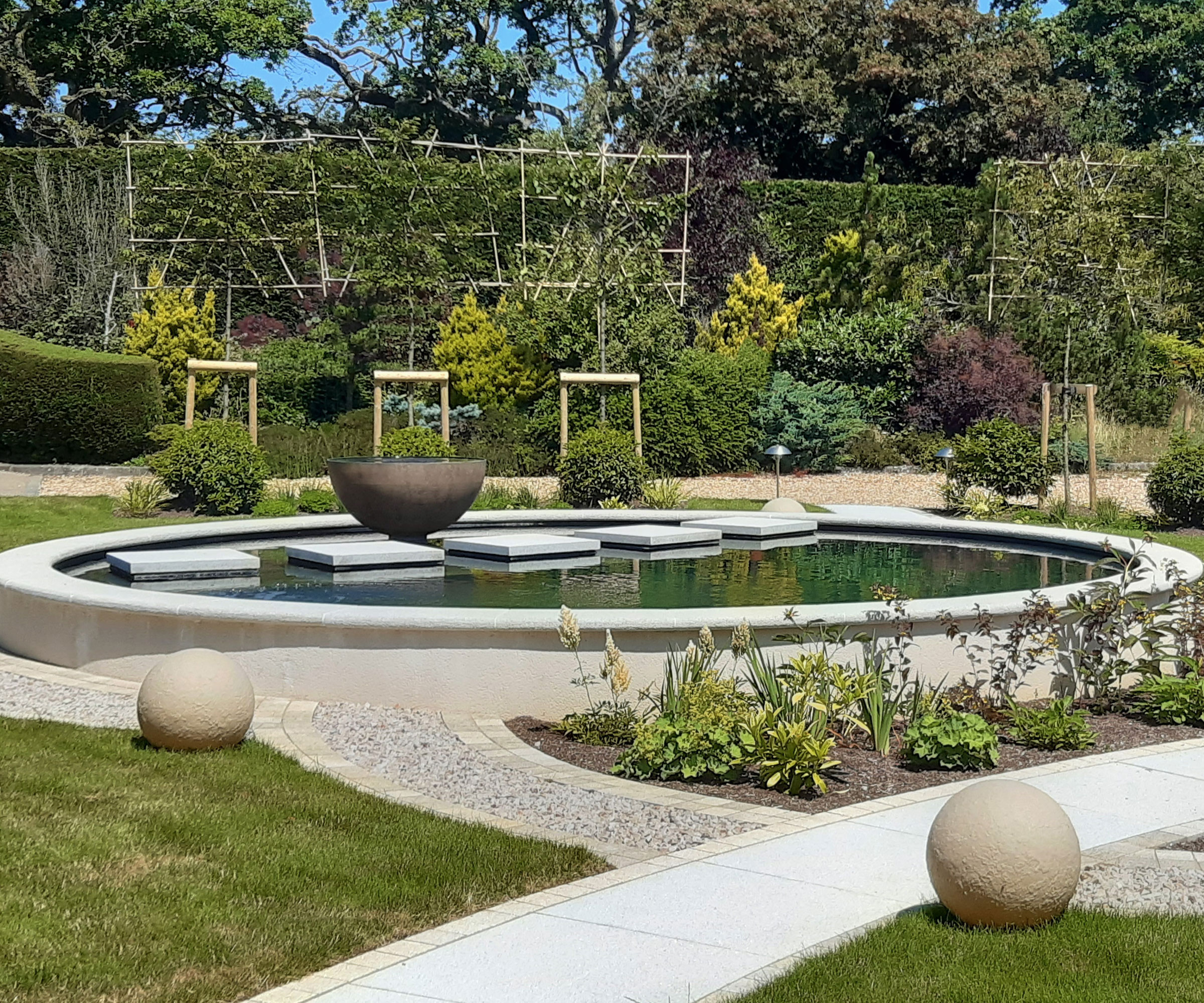 modern circular pond with stepping stone path across it, garden sculptures and intersecting paths, with trees and plants in the background