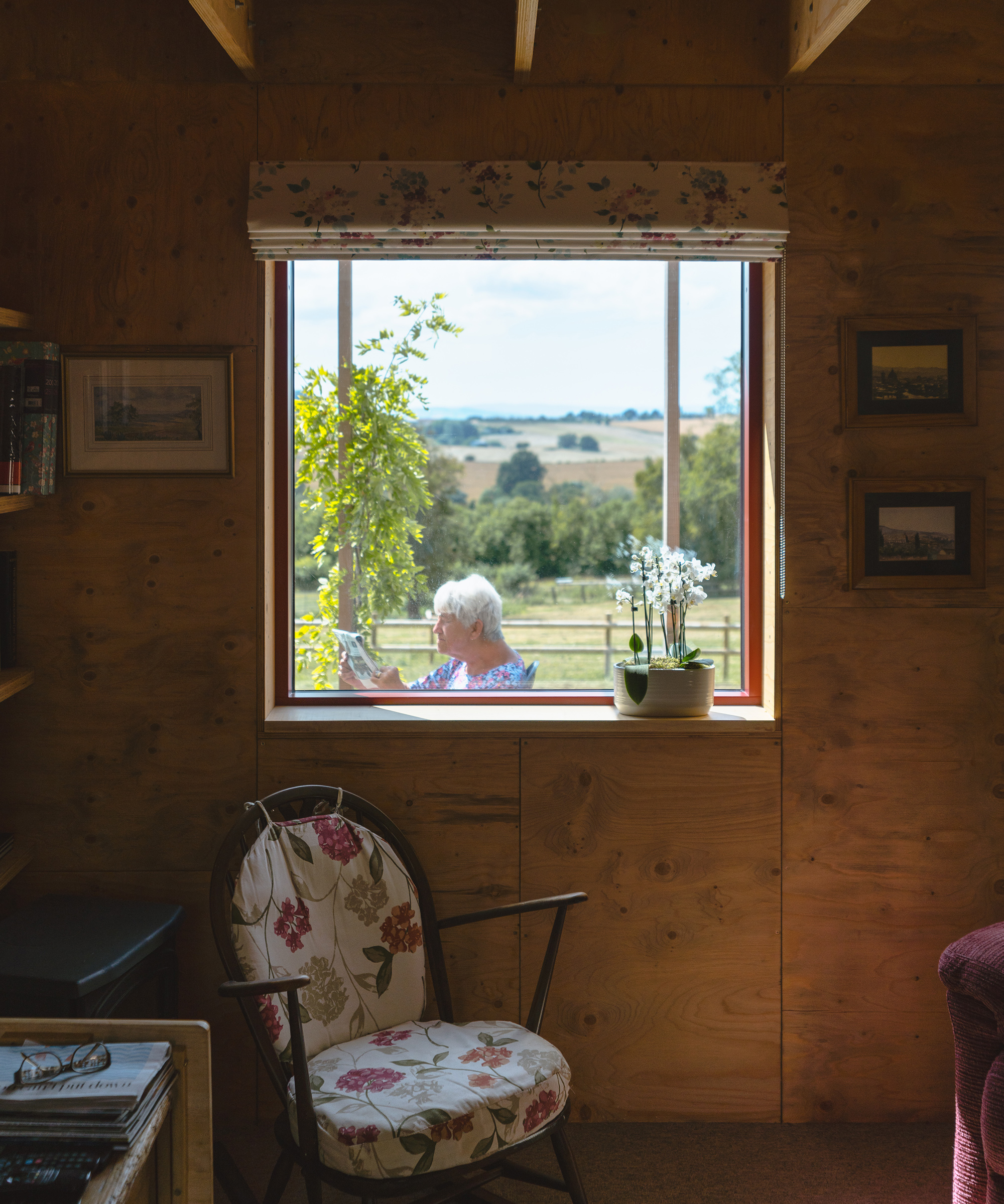 A picture window looking out to a woman sitting outsife on a veranda. The view. isfrom the inside of a living room with timber walls and ceilings
